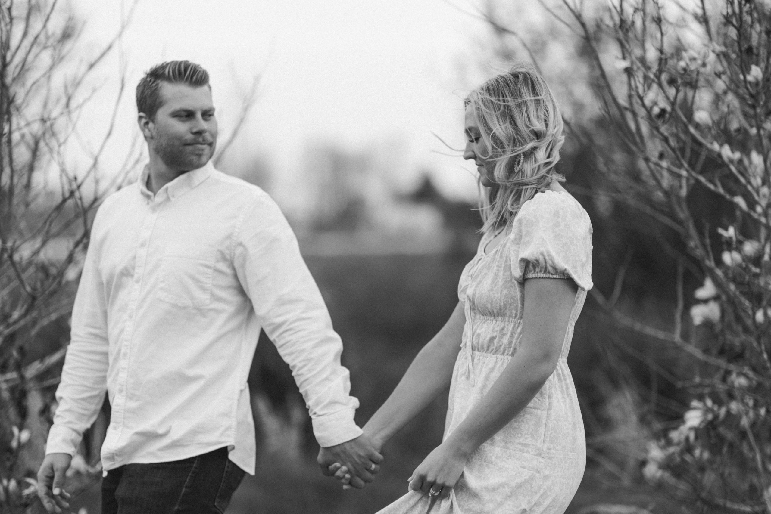 Black and white photo of guy looking back at his fiance as they walk together for their spring engagement photos