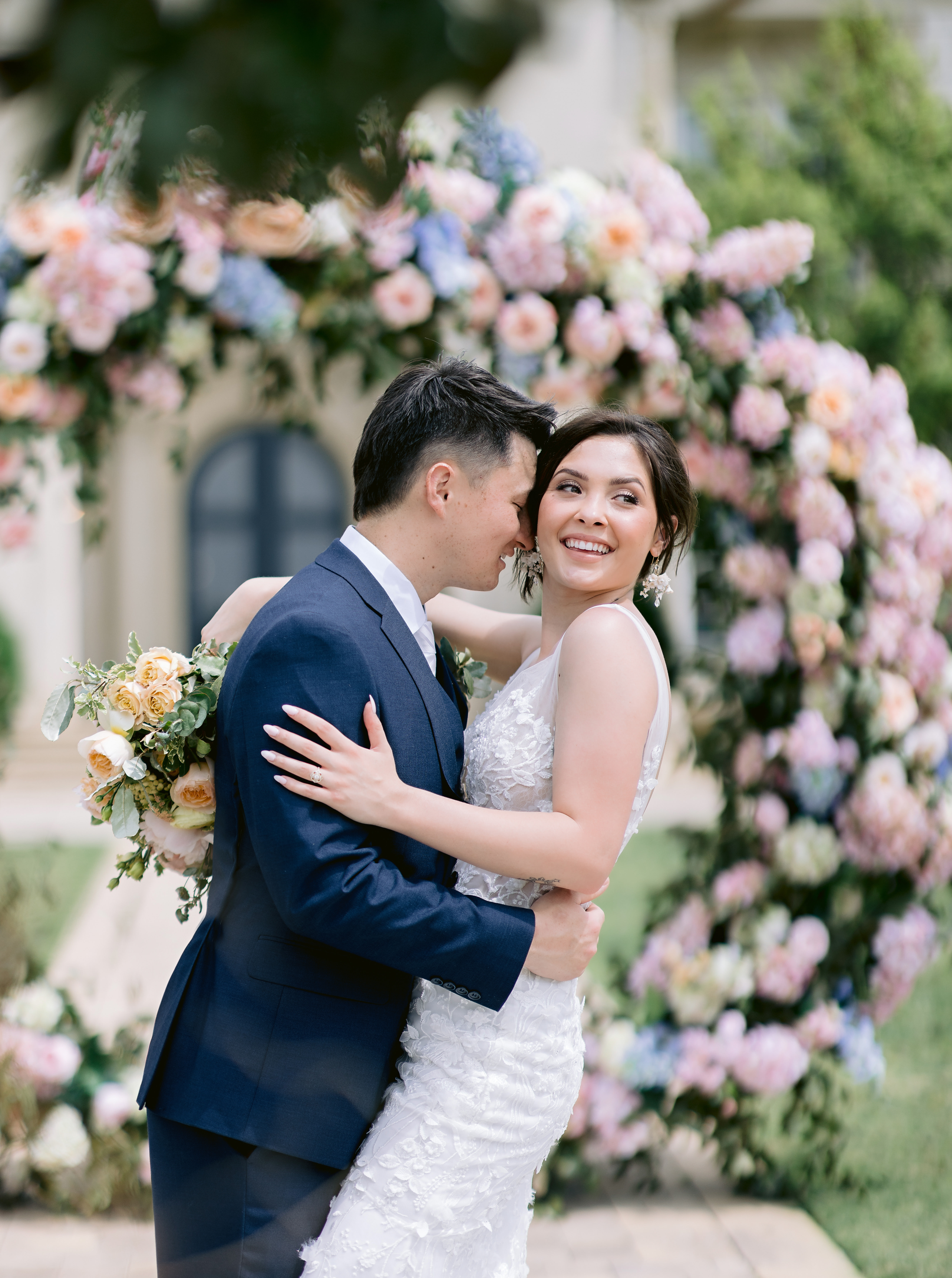 Bride smiles and looks off to the side while groom hugs her in front of the flower arch, captured by Tatyana Zadorin Photography