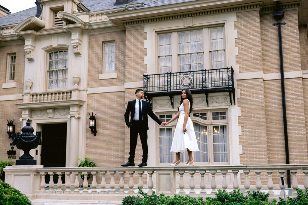 Wide engagement portrait of a couple standing on the balcony outside the historic Aldredge House mansion in Dallas.