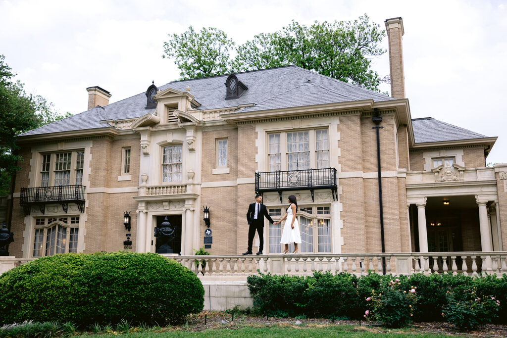 Elegant architectural engagement photo featuring a couple standing in front of the historic Aldredge House surrounded by manicured gardens.