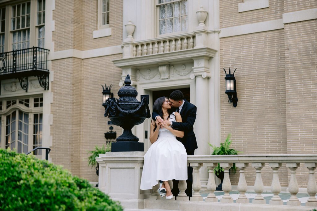 Elegant engagement photo of a couple sharing a kiss in front of the grand entrance to the Aldredge House in Dallas.