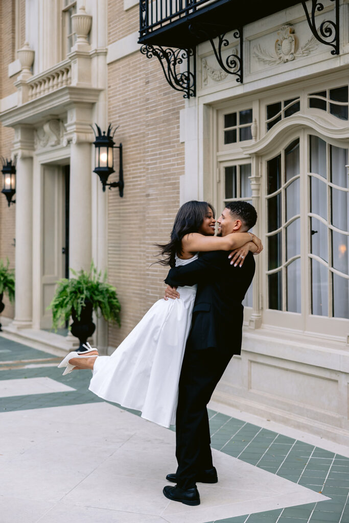 Joyful engagement portrait of a couple embracing as the bride-to-be lifts her leg in front of the historic Aldredge House in Dallas.