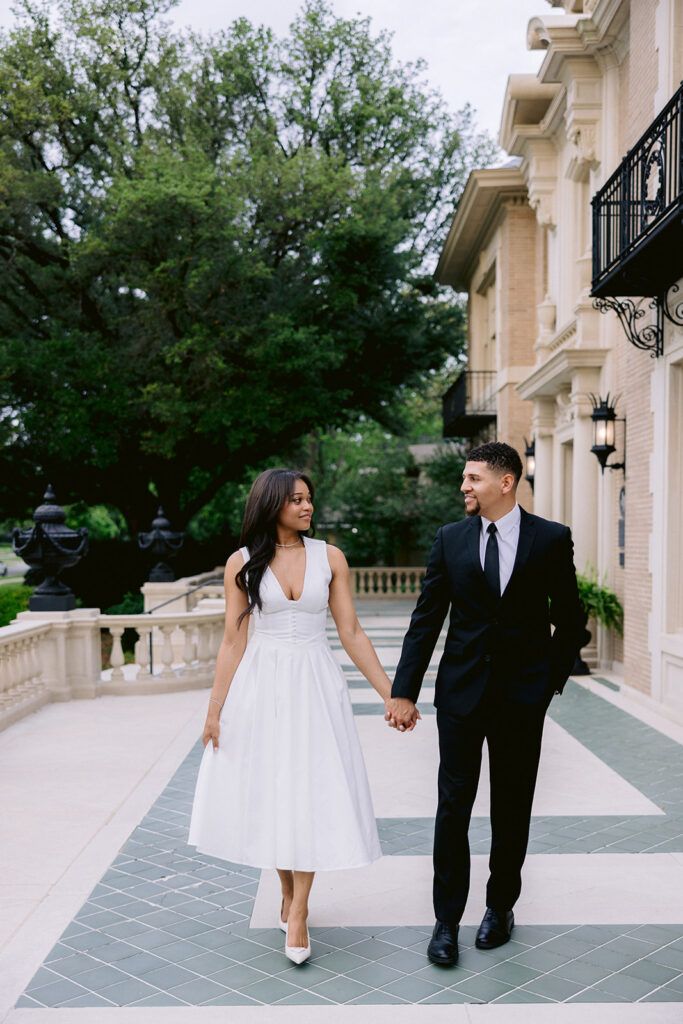 Natural engagement portrait of a couple holding hands while walking beside the Aldredge House mansion in Dallas.