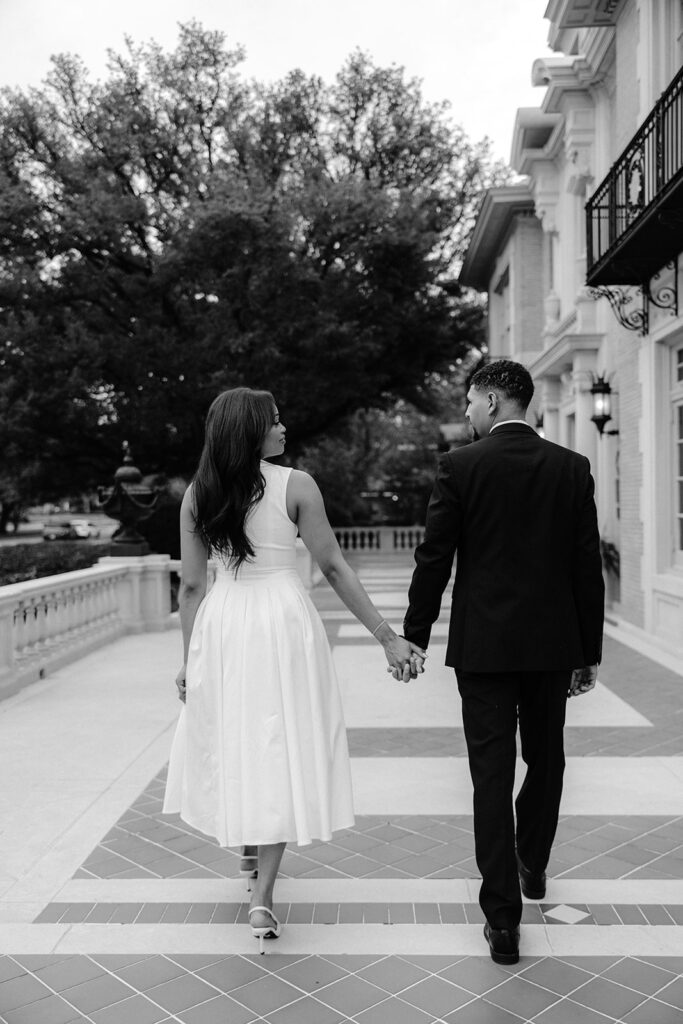 Black-and-white engagement photo of a couple walking away hand in hand through the elegant courtyard at the Aldredge House.