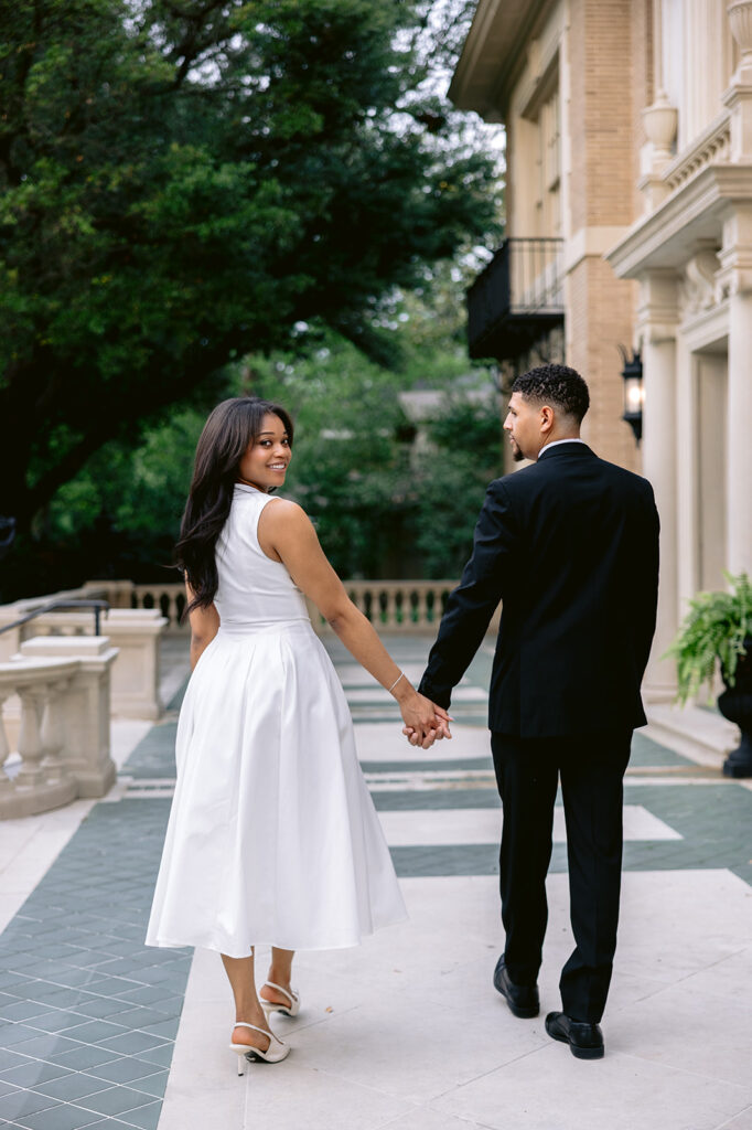 Romantic engagement portrait of a couple looking back while holding hands outside the historic Aldredge House in Dallas.