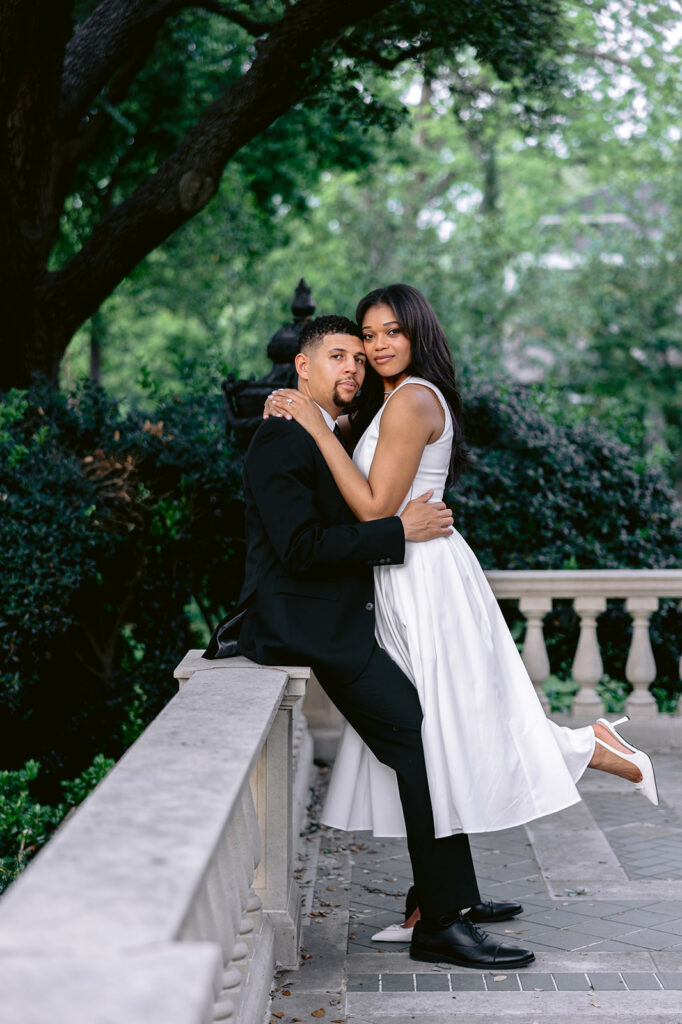 Romantic engagement portrait of a couple embracing on a stone terrace beneath lush trees at the Aldredge House in Dallas, Texas.