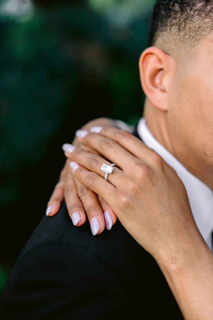 Close-up engagement photo highlighting the bride-to-be's diamond engagement ring resting on her fiancé's shoulder.