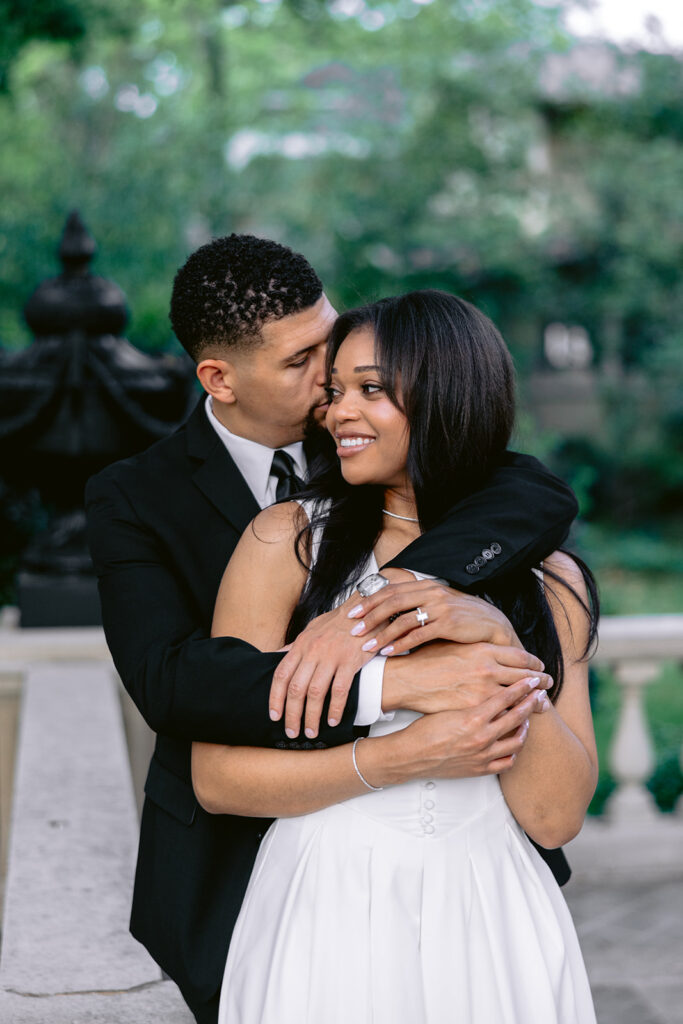 Joyful engagement portrait of a groom hugging his fiancée from behind in the gardens of the Aldredge House in Dallas.