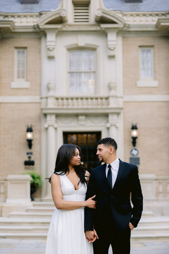 Elegant engagement portrait of a couple smiling at each other in front of the historic Aldredge House in Dallas, Texas.