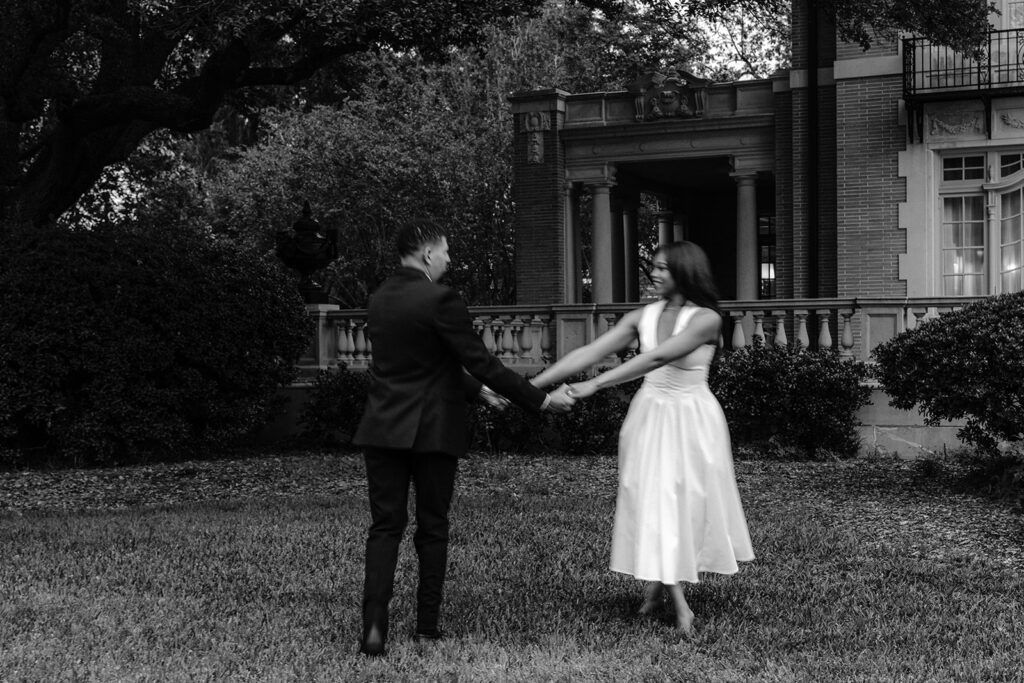 Timeless black-and-white engagement photo of a couple holding hands and spinning together on the lawn at the Aldredge House.