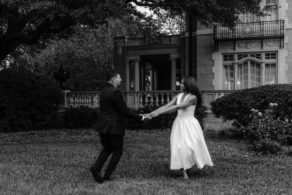 Black-and-white engagement portrait of a couple laughing while holding hands in the gardens of the Aldredge House.