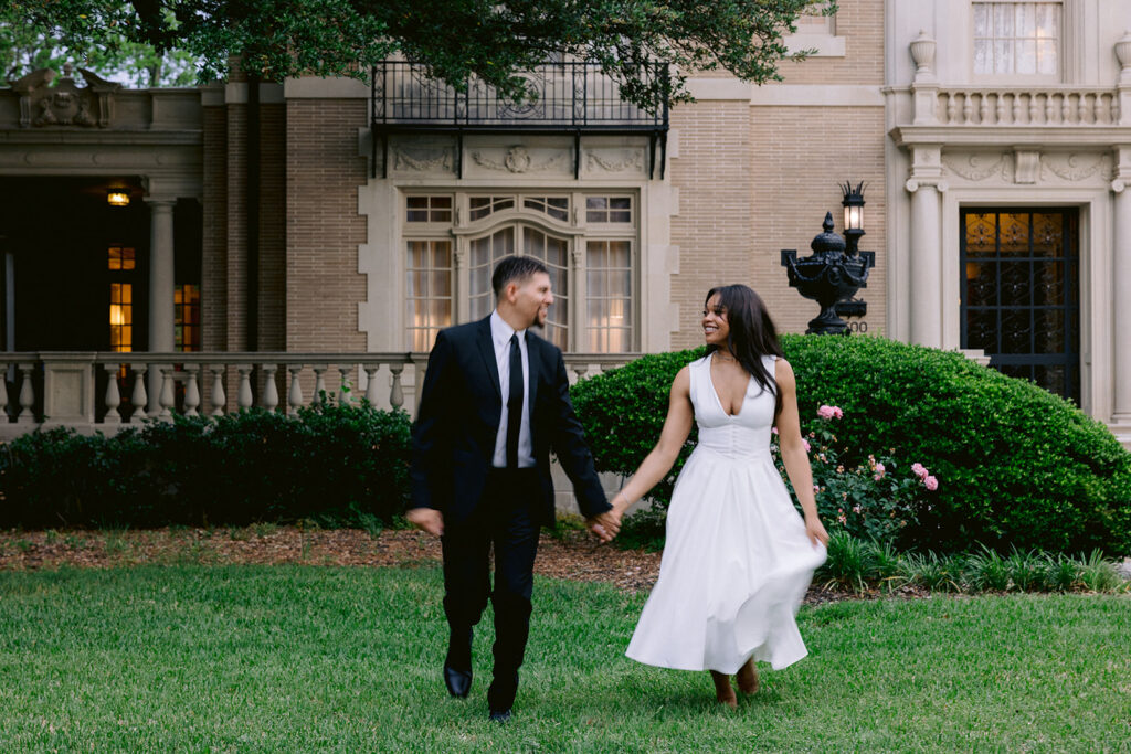 Elegant engagement photo of a couple holding hands and smiling while walking in front of the Aldredge House mansion in Dallas.