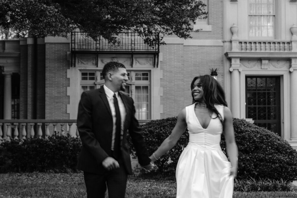 Timeless black-and-white engagement portrait of a couple walking hand in hand outside the historic Aldredge House in Dallas, Texas.