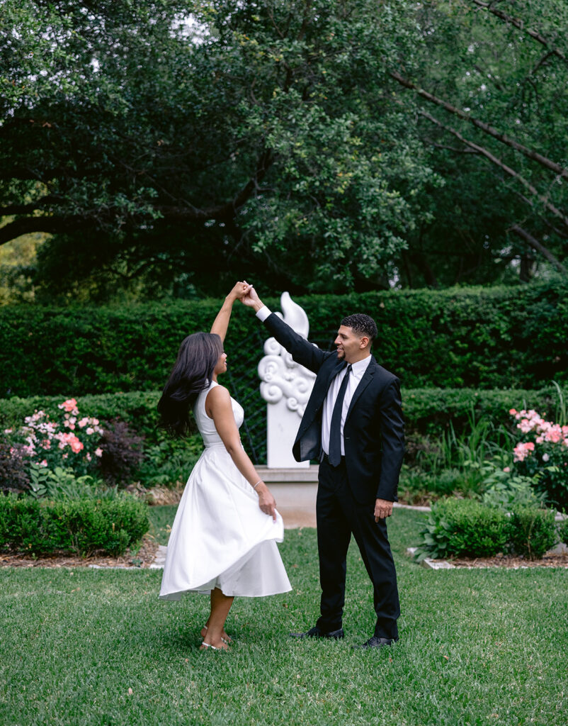Romantic engagement portraits of a couple holding hands and smiling in a lush Dallas garden.
Joyful bride-to-be twirling as her fiancé lifts her hand during an outdoor engagement session at the Aldredge House.
