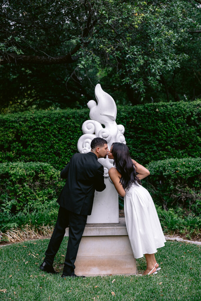 Elegant engagement portrait of a couple embracing beside a white garden sculpture at the Aldredge House in Dallas.