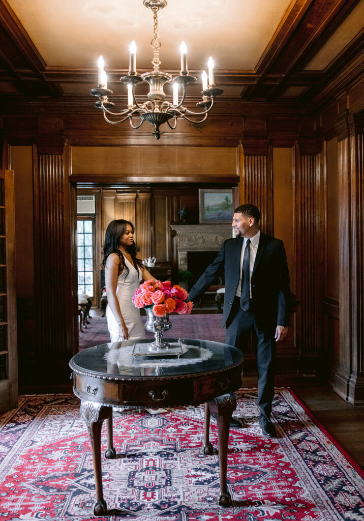 Editorial engagement photo of a couple standing together in a grand historic room featuring elegant wood paneling and a chandelier at the Aldredge House.