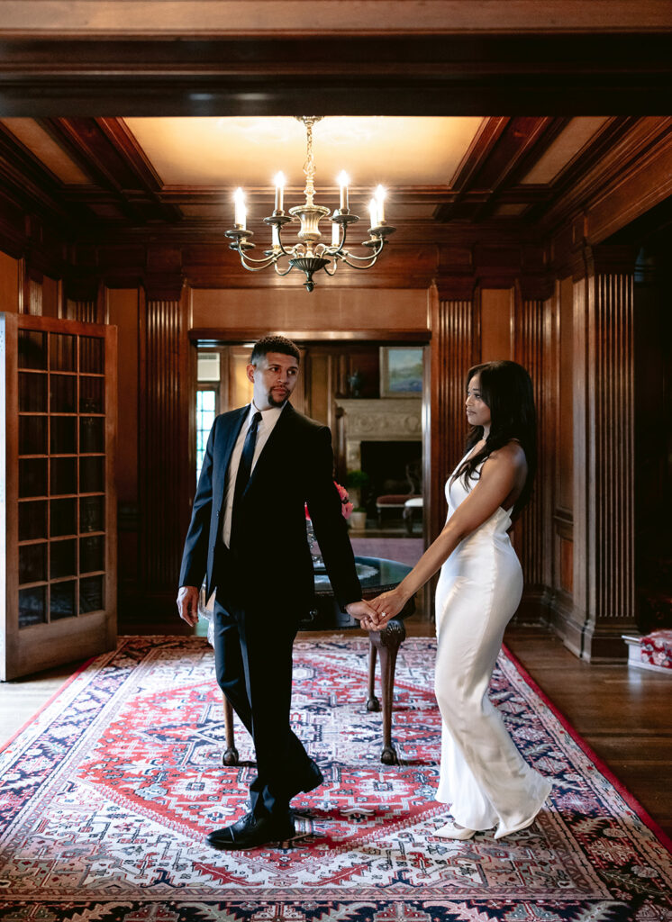 Romantic engagement photo of a couple strolling beneath a crystal chandelier inside the historic Aldredge House mansion.