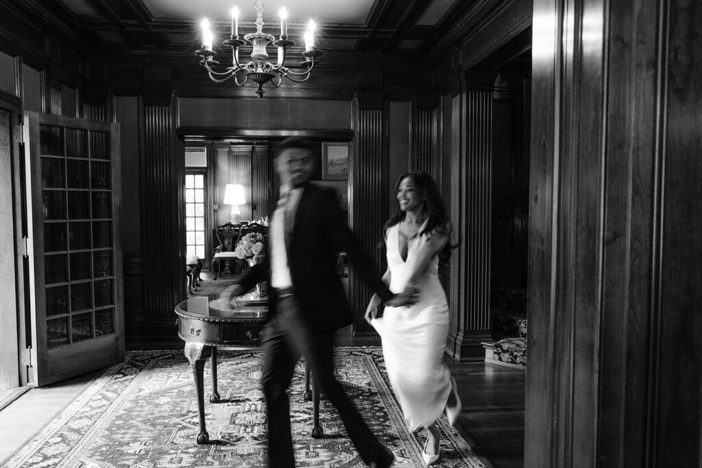 Timeless black-and-white engagement photo of a couple sharing a romantic walk through the elegant interior of the Aldredge House.