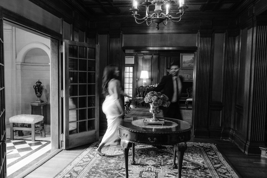 Timeless black-and-white engagement portrait of a couple walking through the elegant historic interior of the Aldredge House in Dallas, Texas.