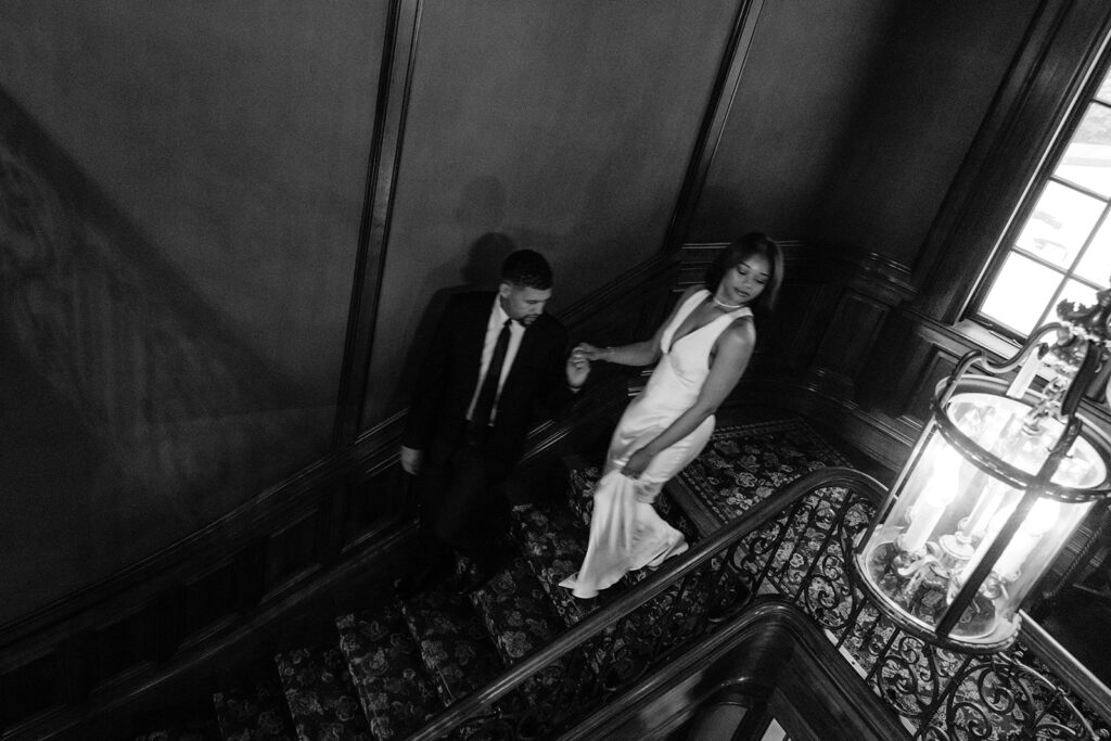 Black-and-white engagement portrait of a couple descending the elegant staircase during their luxury indoor engagement session.