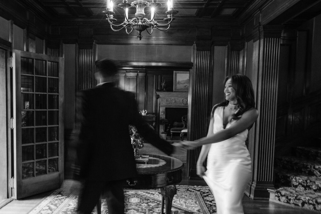 Black-and-white engagement portrait of a couple holding hands while walking through the elegant Aldredge House in Dallas.