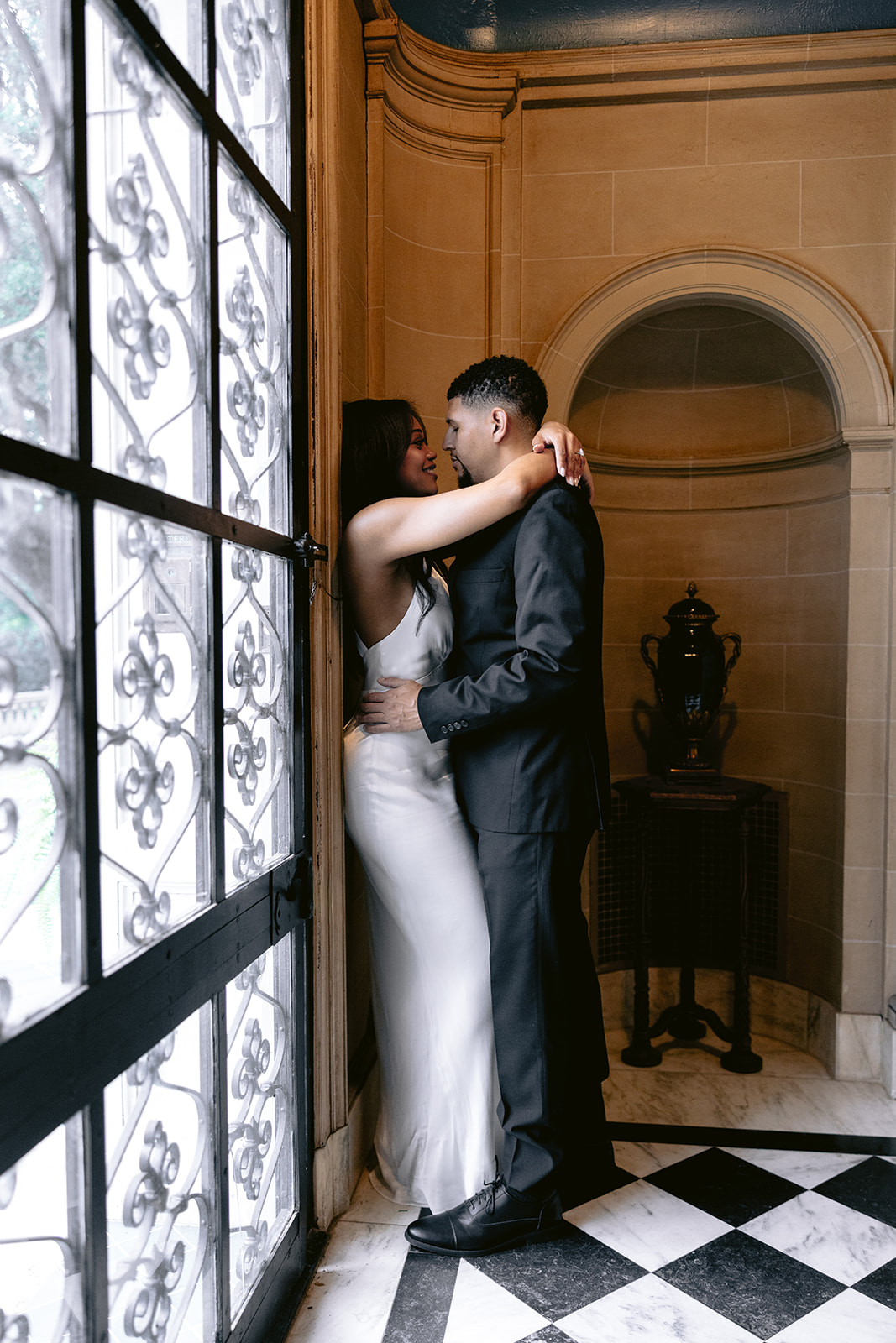 Editorial engagement photo of a couple embracing beside an ornate leaded glass window inside the Aldredge House mansion.