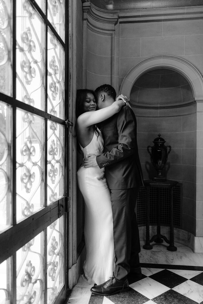 Black-and-white engagement portrait of a couple embracing by an ornate window inside the Aldredge House mansion.