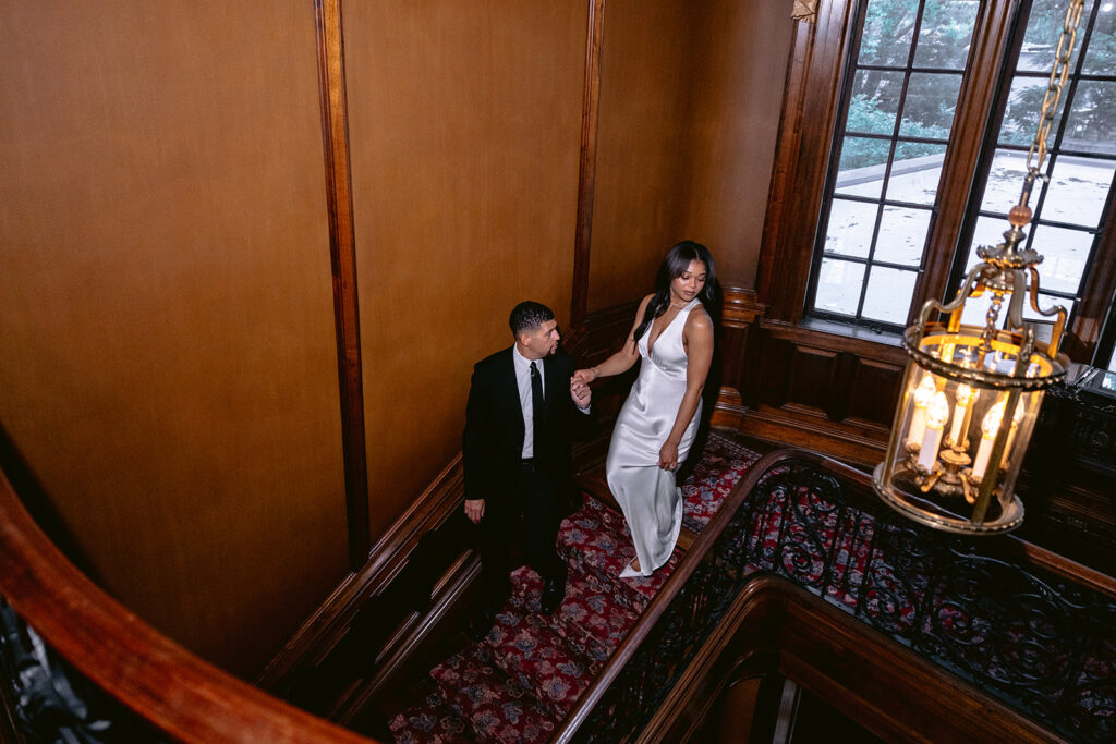 Romantic engagement photo of a couple walking hand in hand down the grand staircase beneath a historic chandelier at the Aldredge House.