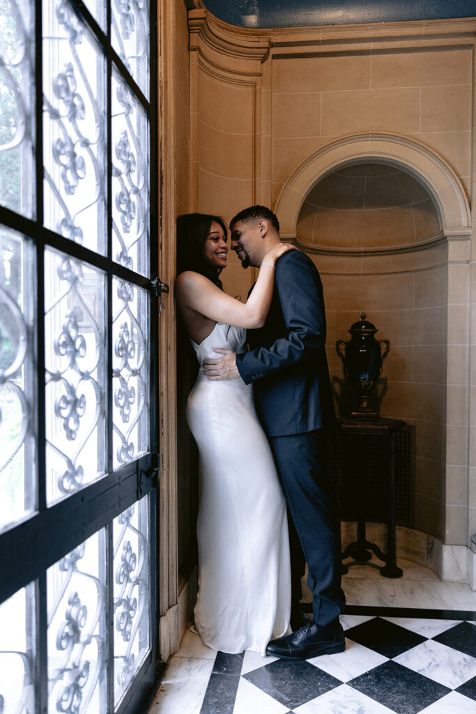 Romantic engagement portrait of a couple embracing beside an ornate window inside the historic Aldredge House in Dallas, Texas.