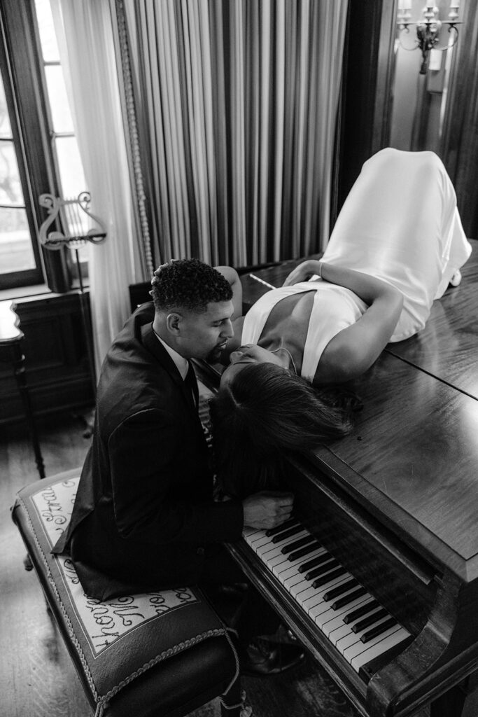 Timeless black-and-white engagement photo of a groom seated at a grand piano while his fiancée leans over him inside the Aldredge House.