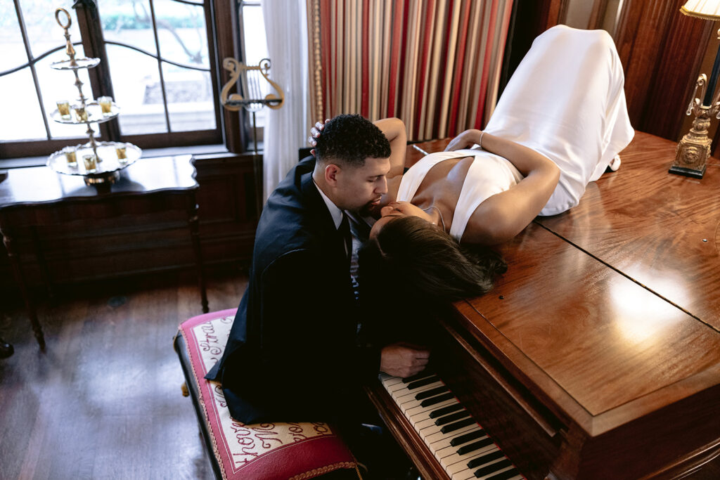 Editorial engagement portrait of a couple sharing a romantic kiss beside a grand piano in the historic Aldredge House mansion.