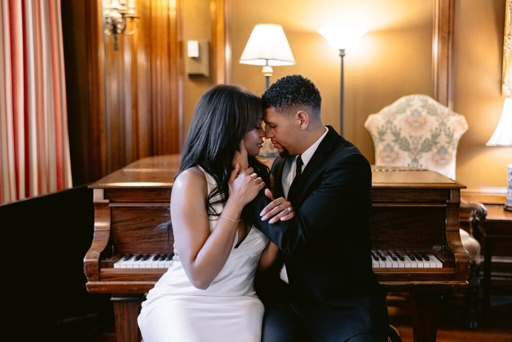 Romantic engagement portrait of a couple sharing a kiss while seated beside a grand piano inside the historic Aldredge House mansion.