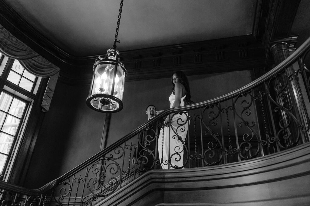 Editorial engagement portrait of a bride-to-be standing on the grand staircase beneath a historic chandelier inside the Aldredge House.