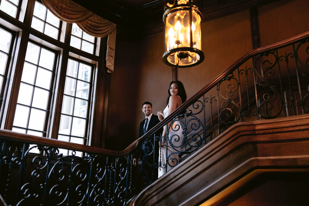 Elegant engagement photo of a couple on the grand staircase beneath a vintage chandelier in the historic Aldredge House mansion.