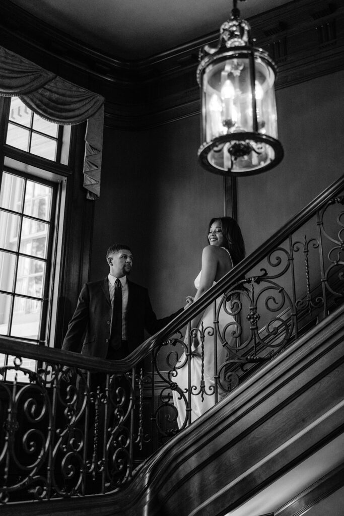 Timeless black-and-white engagement portrait of a couple standing together on the ornate staircase inside the Aldredge House in Dallas, Texas.