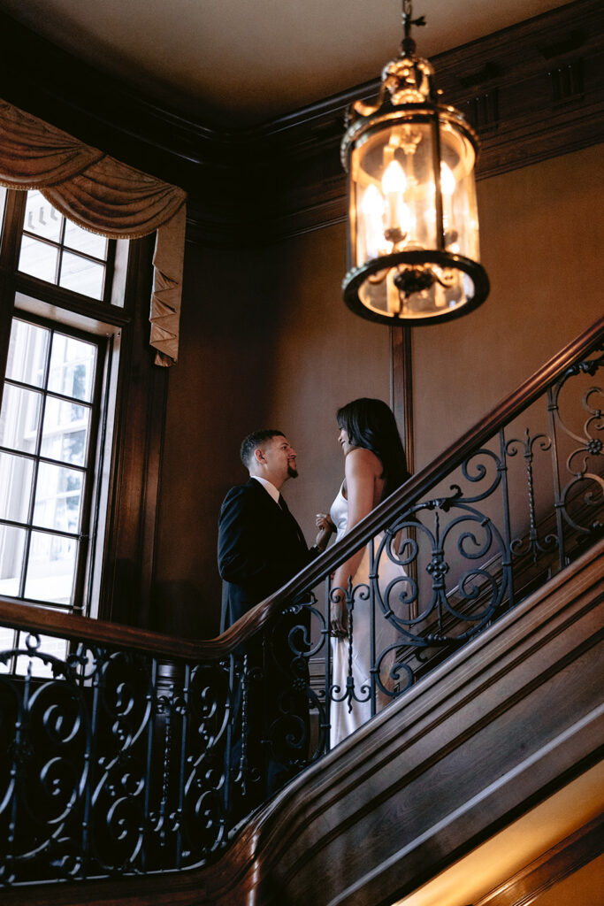 Luxury engagement portrait of a couple sharing a romantic moment on the grand staircase beneath an elegant chandelier at the Aldredge House.