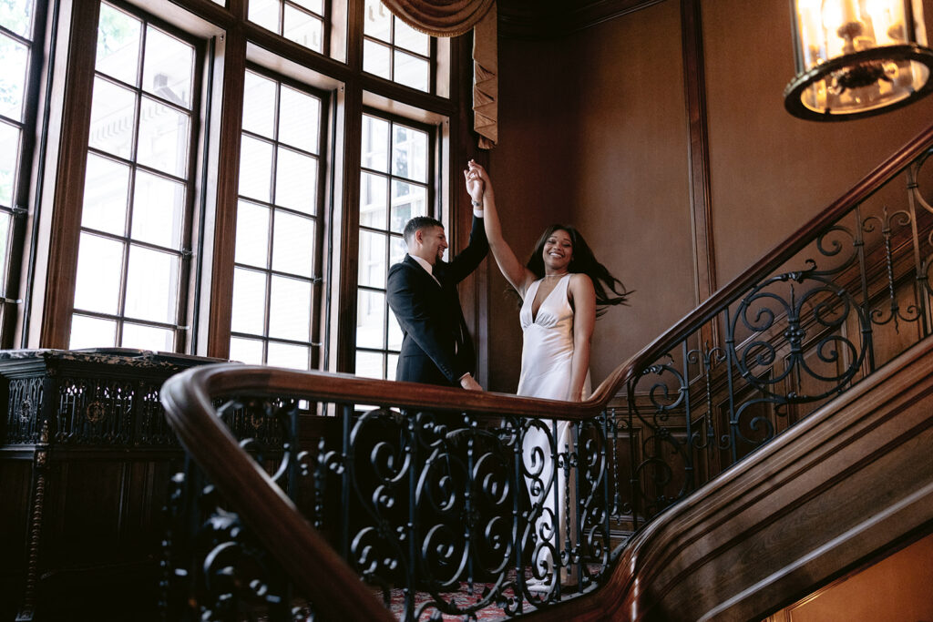 Elegant engagement photo of a couple holding hands on the ornate staircase beneath a vintage chandelier at the Aldredge House.