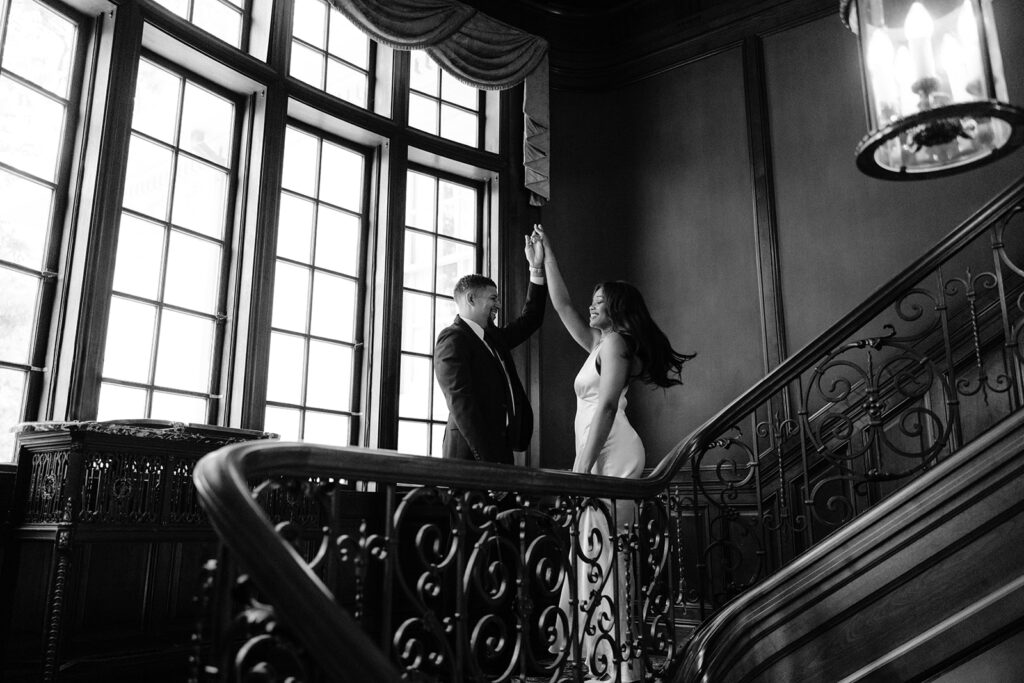 Timeless black-and-white engagement portrait of a couple standing together on the grand staircase inside the Aldredge House mansion.