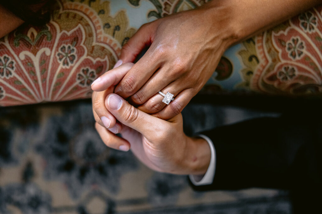 Natural engagement photo of a smiling bride-to-be showing her engagement ring while sharing an intimate moment with her fiancé indoors.