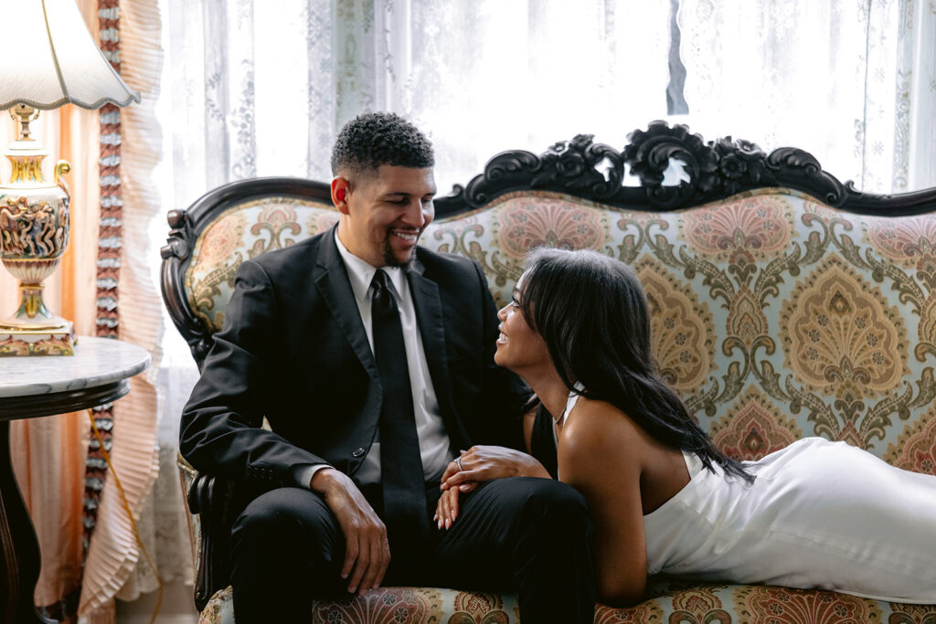 Romantic indoor engagement photo of a couple smiling at each other while seated on an antique sofa in the Aldredge House.