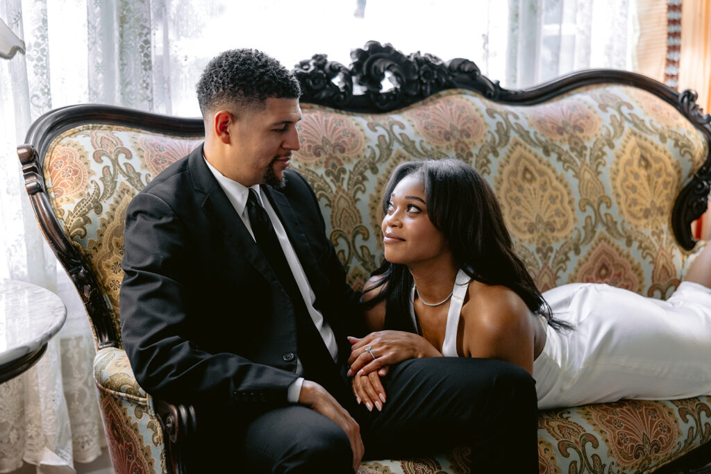 Editorial engagement portrait of a groom admiring his fiancée as they relax together in an elegant historic mansion.