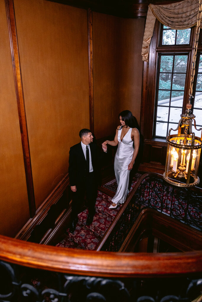 Luxury engagement photo of a couple standing together on the grand staircase surrounded by warm wood architecture and timeless interiors.