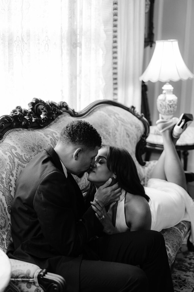 Black-and-white close-up engagement photo of a couple sharing a kiss on an antique sofa inside the Aldredge House.
