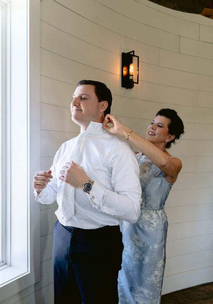 Groom getting ready for his wedding day with candid moments adjusting his tie, putting on his suit jacket, sharing a heartfelt hug with his mother, and elegant portraits at The French Farmhouse wedding venue in Collinsville, Texas.