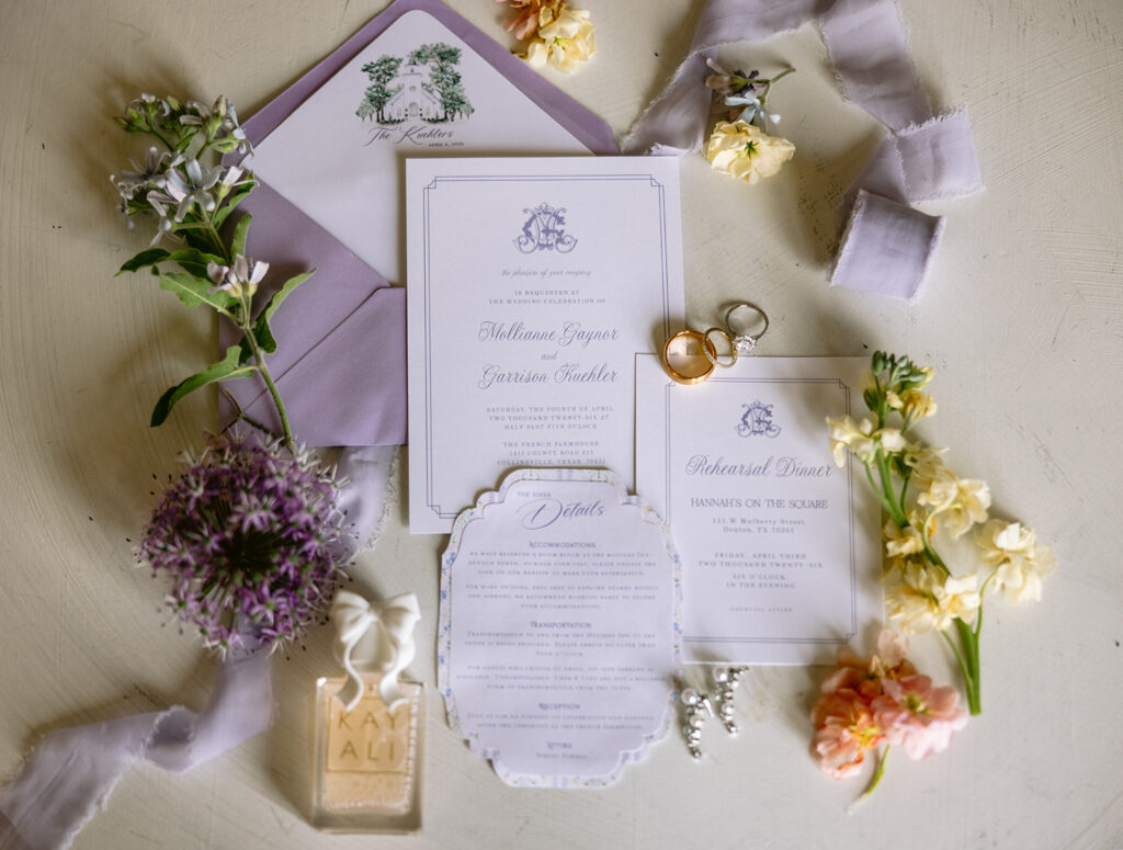 Flat lay wedding details featuring elegant invitation suite, gold wedding rings, vow books, bridal shoes, and fine styling with soft florals at The French Farmhouse wedding venue in Collinsville, Texas