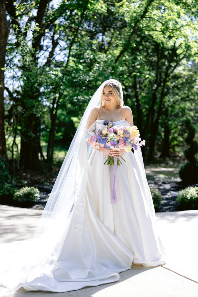 Romantic bridal portraits featuring the bride in a timeless off-the-shoulder wedding gown with a flowing cathedral veil, holding a colorful pastel spring bouquet during elegant outdoor portraits at The French Farmhouse wedding venue in Collinsville, Texas.