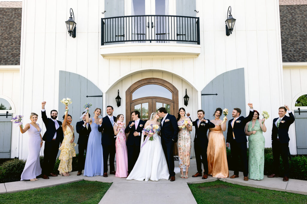 Elegant wedding party portraits featuring the bride and groom with bridesmaids in colorful pastel dresses and groomsmen in classic black suits, celebrating together outside The French Farmhouse wedding venue in Collinsville, Texas.