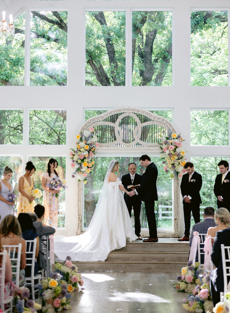 Bright chapel wedding ceremony featuring the bride and groom exchanging vows, colorful spring floral arrangements, bridesmaids in pastel dresses, and elegant ceremony moments at The French Farmhouse wedding venue in Collinsville, Texas.