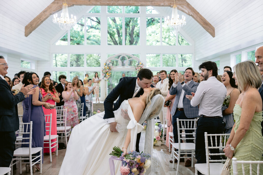 Joyful wedding ceremony recessional featuring the newly married bride and groom walking down the aisle, sharing their first kiss as husband and wife, and celebrating with cheering family and friends at The French Farmhouse wedding venue in Collinsville, Texas.