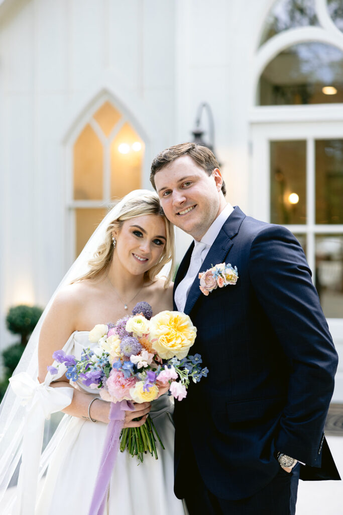 Romantic bride and groom sunset portraits outside the white chapel at The French Farmhouse wedding venue in Collinsville, Texas, featuring a colorful pastel bouquet, timeless wedding attire, and elegant newlywed moments.